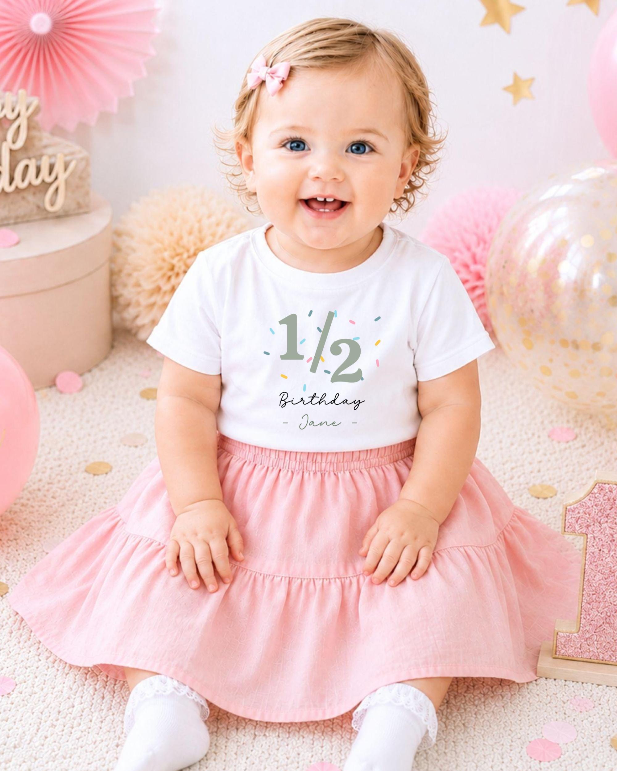A smiling baby girl wearing a white t-shirt with the number 1/2 and a pink skirt, sitting on a pink and white background with balloons and confetti.