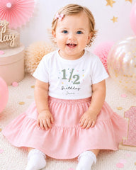A smiling baby girl wearing a white t-shirt with the number 1/2 and a pink skirt, sitting on a pink and white background with balloons and confetti.