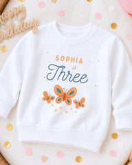 A white sweatshirt with a personalized design, featuring the text "Sophia is Three" and three butterflies. The sweatshirt is placed on a surface with confetti and a wooden sign that says "Happy Birthday".