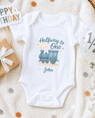 A white onesie with a train graphic and the text "Halfway to One John" printed on it, along with a "Happy Birthday" sign and a gift box.