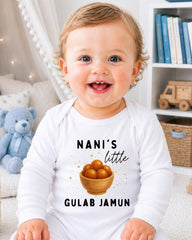 A smiling baby wearing a white onesie with a graphic and text printed on it, sitting in a room with a teddy bear and bookshelf in the background.