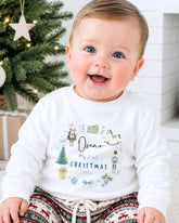A smiling baby boy wearing a white sweatshirt with a personalized design and text, sitting in front of a Christmas tree.