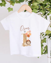 A white t-shirt with a graphic design featuring a lion and balloons, along with the text "Oscar is" printed on it.