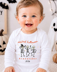 A smiling baby wearing a white onesie with a Halloween-themed design, including three cartoon skeletons and the text "my first Halloween Alexander 20.26".