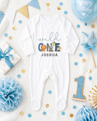 A white onesie with the text "wild one Joshua" printed on it, surrounded by various baby-themed items such as a blue balloon, a blue and gold star, and a blue and gold bow.
