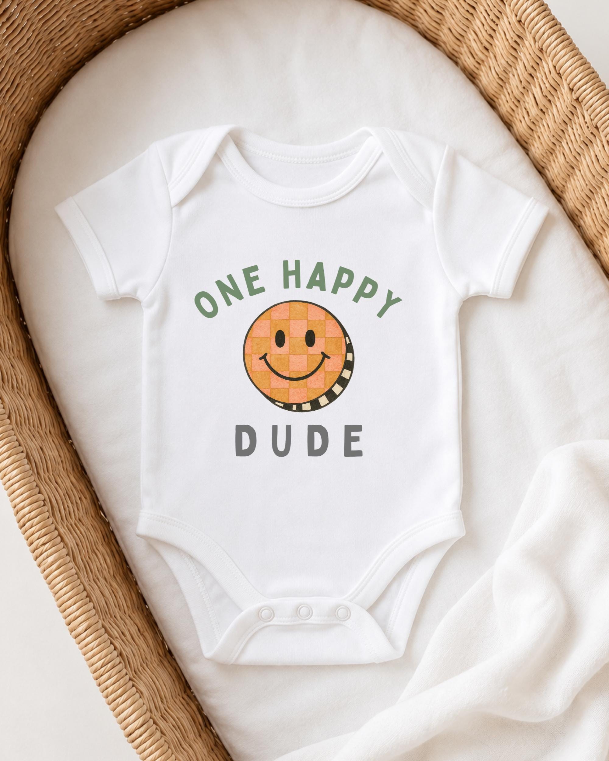 A white onesie with a smiley face and the text "ONE HAPPY DUDE" printed on it, laid out on a white blanket.