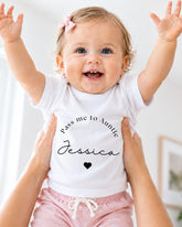 A smiling baby girl wearing a white t-shirt with the text "Pass me to Aunt Jessica" and pink shorts, being held up by someone's arms.