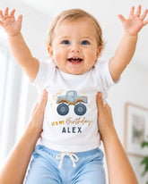 A smiling baby wearing a white t-shirt with a truck graphic and the text "It's my birthday Alex" is being held up by someone's arms.