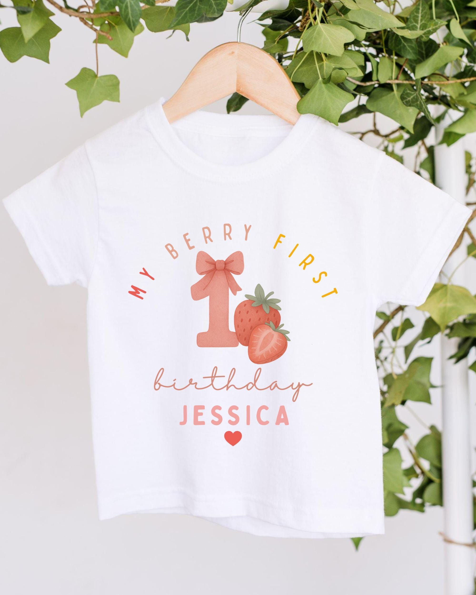 A white t-shirt with a graphic design featuring a pink number one, strawberries, and the text "My Berry First birthday Jessica" printed on it.