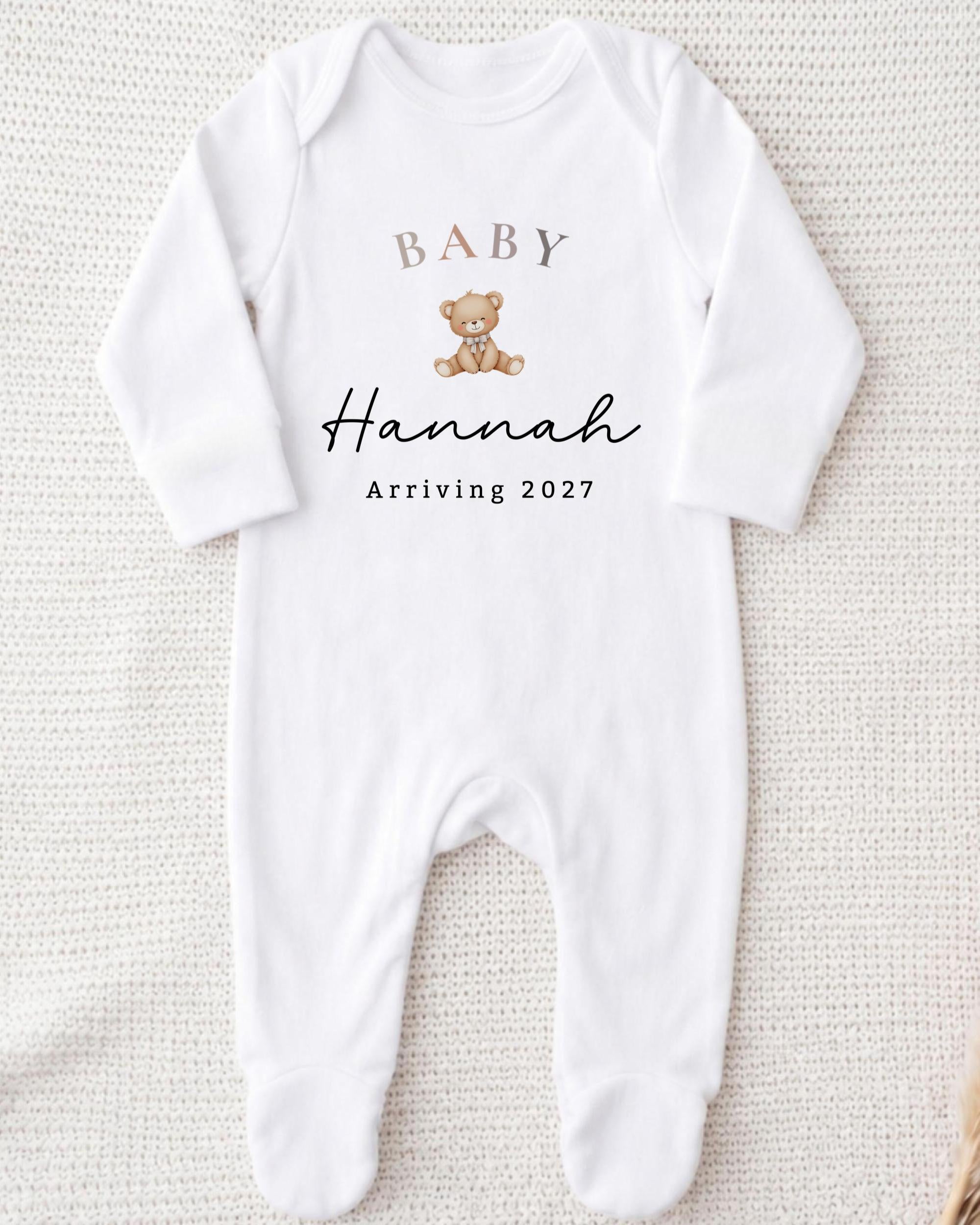 A white onesie with a teddy bear and text that says "BABY Hannah arriving 2027".