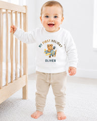 A smiling baby boy wearing a white sweatshirt with a cartoon image of a bear and the text "My first holiday Oliver" stands next to a wooden crib.