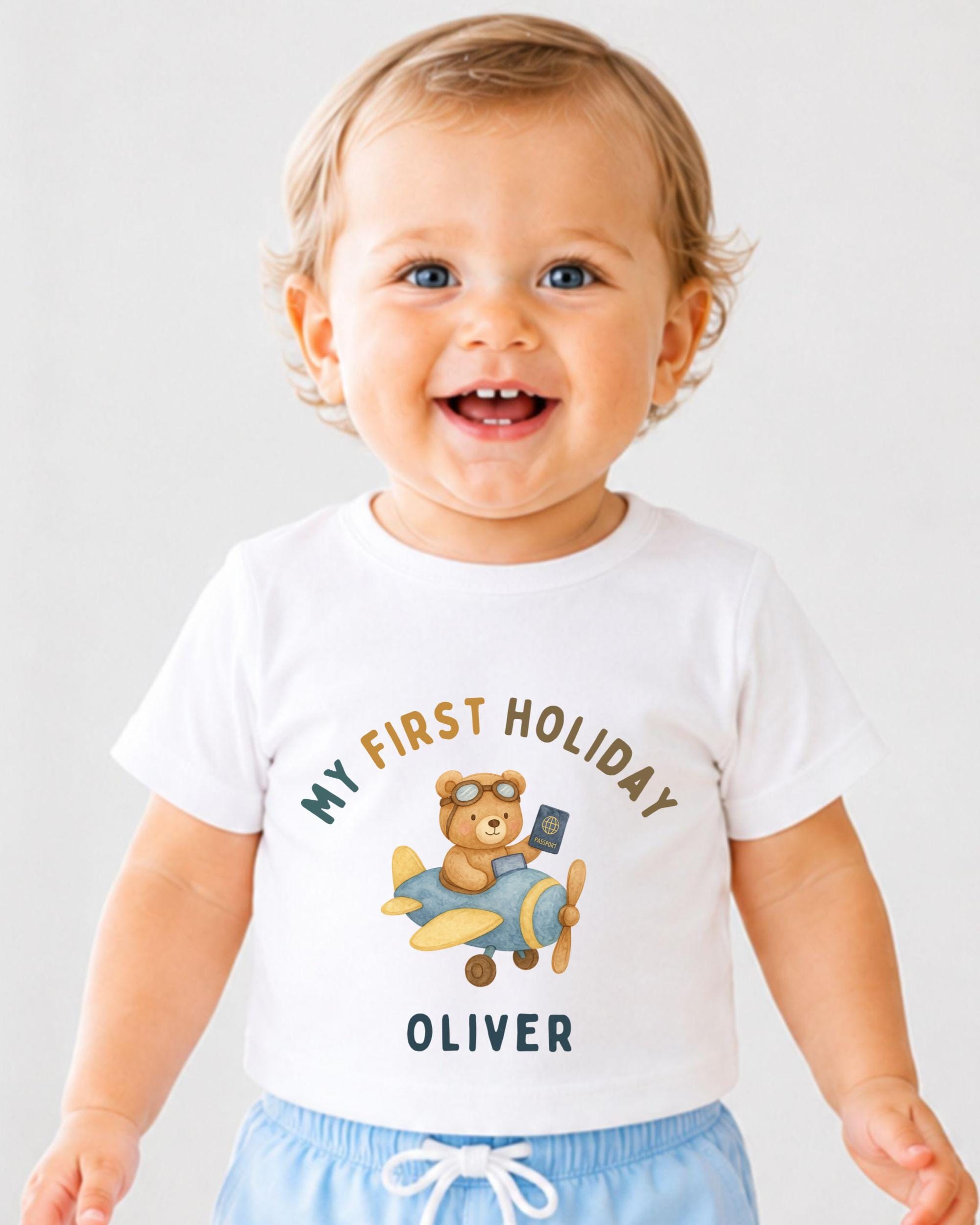 A smiling baby boy wearing a white t-shirt with a graphic of a teddy bear and an airplane, along with the text "My first holiday Oliver".