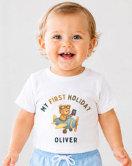 A smiling baby boy wearing a white t-shirt with a graphic of a teddy bear and an airplane, along with the text "My first holiday Oliver".
