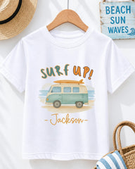 A white t-shirt with a graphic design of a van and the text "Surf up! Jackson" printed on it, along with a surfboard. The t-shirt is hanging on a wooden hanger, and there is a straw hat and a "Beach Sun Waves" sign visible in the background.