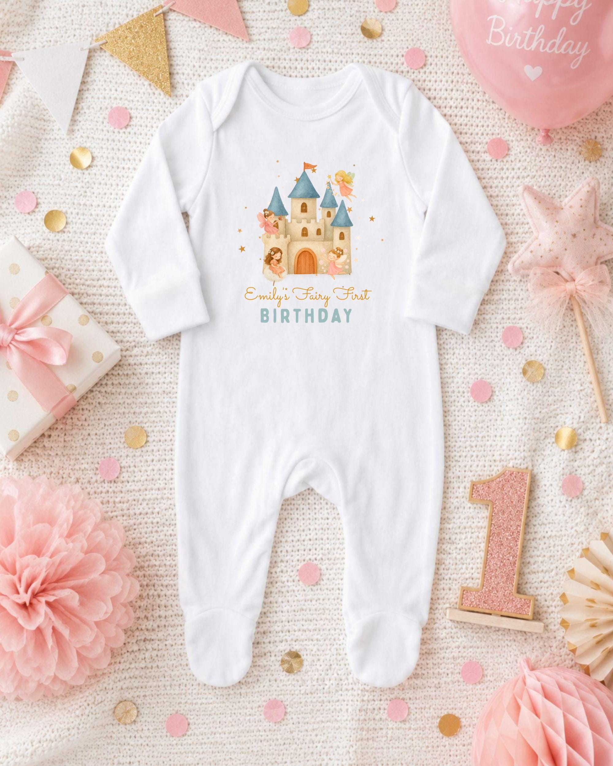 A white onesie with a castle design and the text "Emilly's Fairy Tale Birthday" printed on it, surrounded by pink and gold confetti, a pink star, and a pink and gold birthday cake.