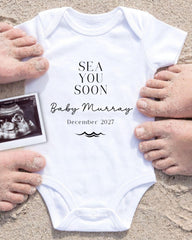 a white onesie with the text "SEA YOU SOON Baby Murray December 2027" printed on it, surrounded by sand and a black and white photograph of a baby's belly.