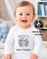 A smiling baby wearing a white onesie with a dog design, sitting in a room with a teddy bear and bookshelf in the background.