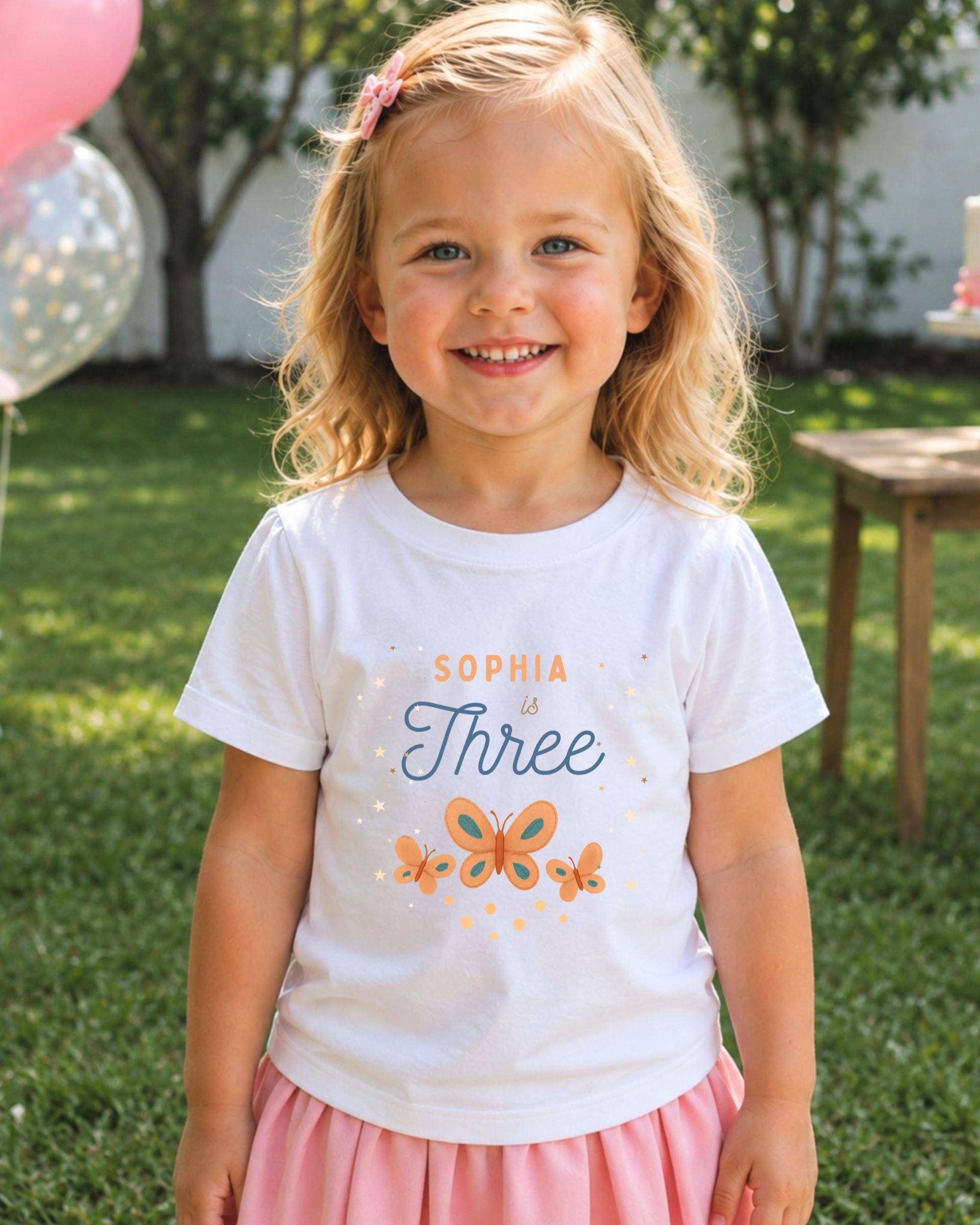 A young girl with blonde hair is smiling and wearing a white t-shirt with a personalized message and butterflies printed on it.