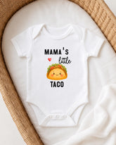 A white onesie with a cartoon taco printed on it, placed on a white blanket.