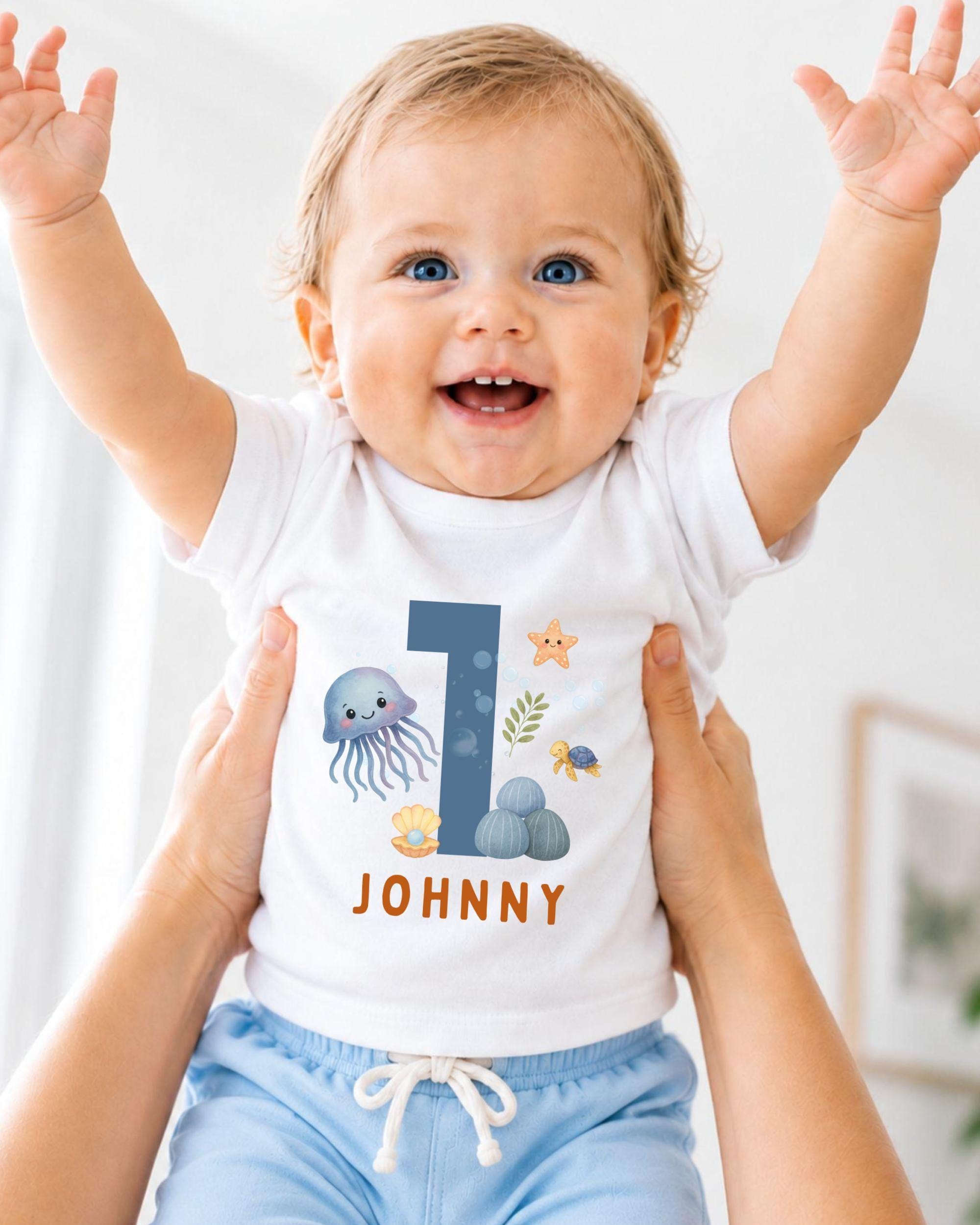 A smiling baby wearing a white onesie with a blue number 1 and ocean-themed designs.
