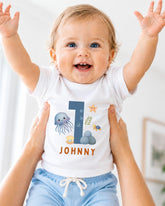 A smiling baby wearing a white bodysuit with a blue number 1 and ocean-themed designs.