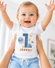 A smiling baby wearing a white onesie with a blue number 1 and ocean-themed designs.