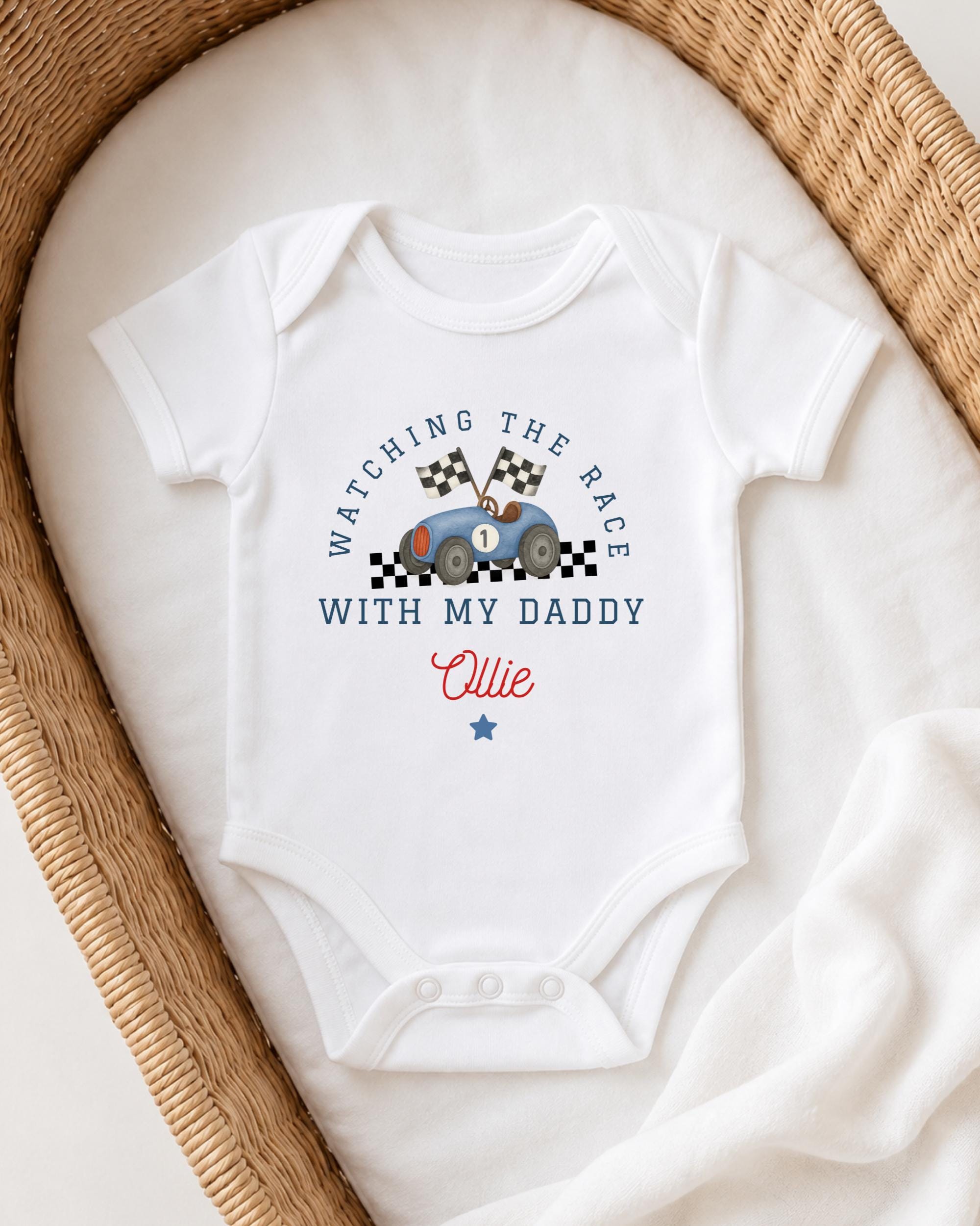 A white onesie with a racing car design and the text "Watching the race with my daddy" printed on it, placed in a woven basket.