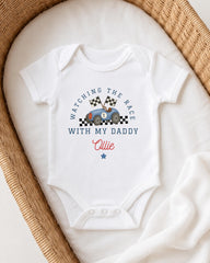 A white onesie with a racing car design and the text "Watching the race with my daddy" printed on it, placed in a woven basket.