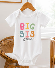A white baby onesie with the text "BIG SIS Jessica" printed on it, hanging on a wooden hanger.
