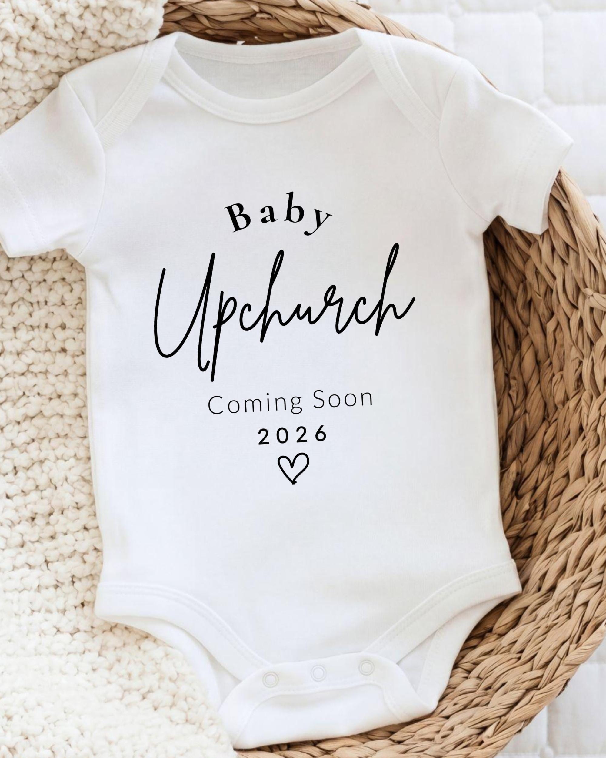 A white baby onesie with the text "Baby Upchurch Coming Soon 2026" printed on it, placed on a woven basket.