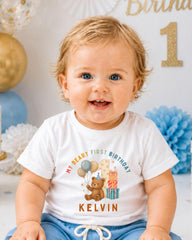 A smiling baby boy wearing a white t-shirt with a teddy bear and birthday decorations, sitting in front of a birthday party backdrop.