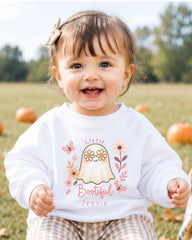 A smiling baby girl wearing a white sweater with a ghost and floral design, sitting in a field with pumpkins in the background.