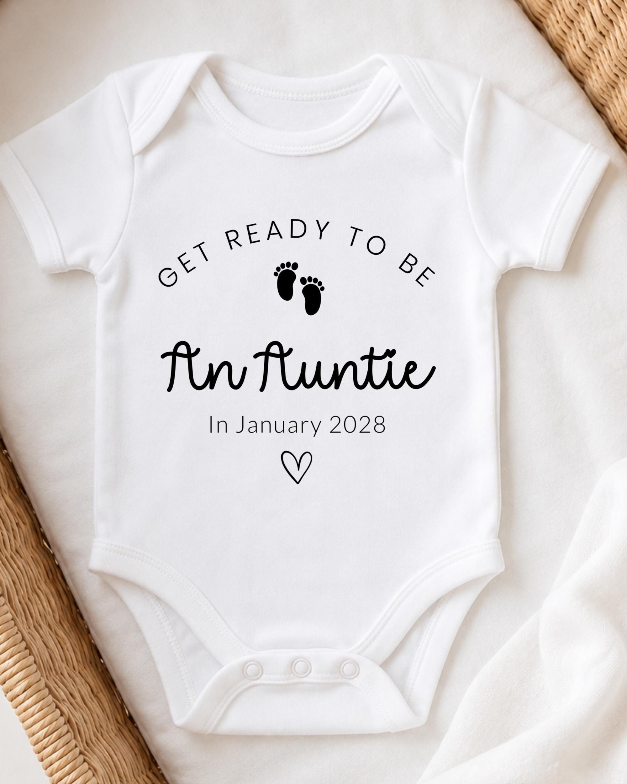 A white baby onesie with the text "Get ready to be An Auntie" printed on it, along with a small heart symbol.