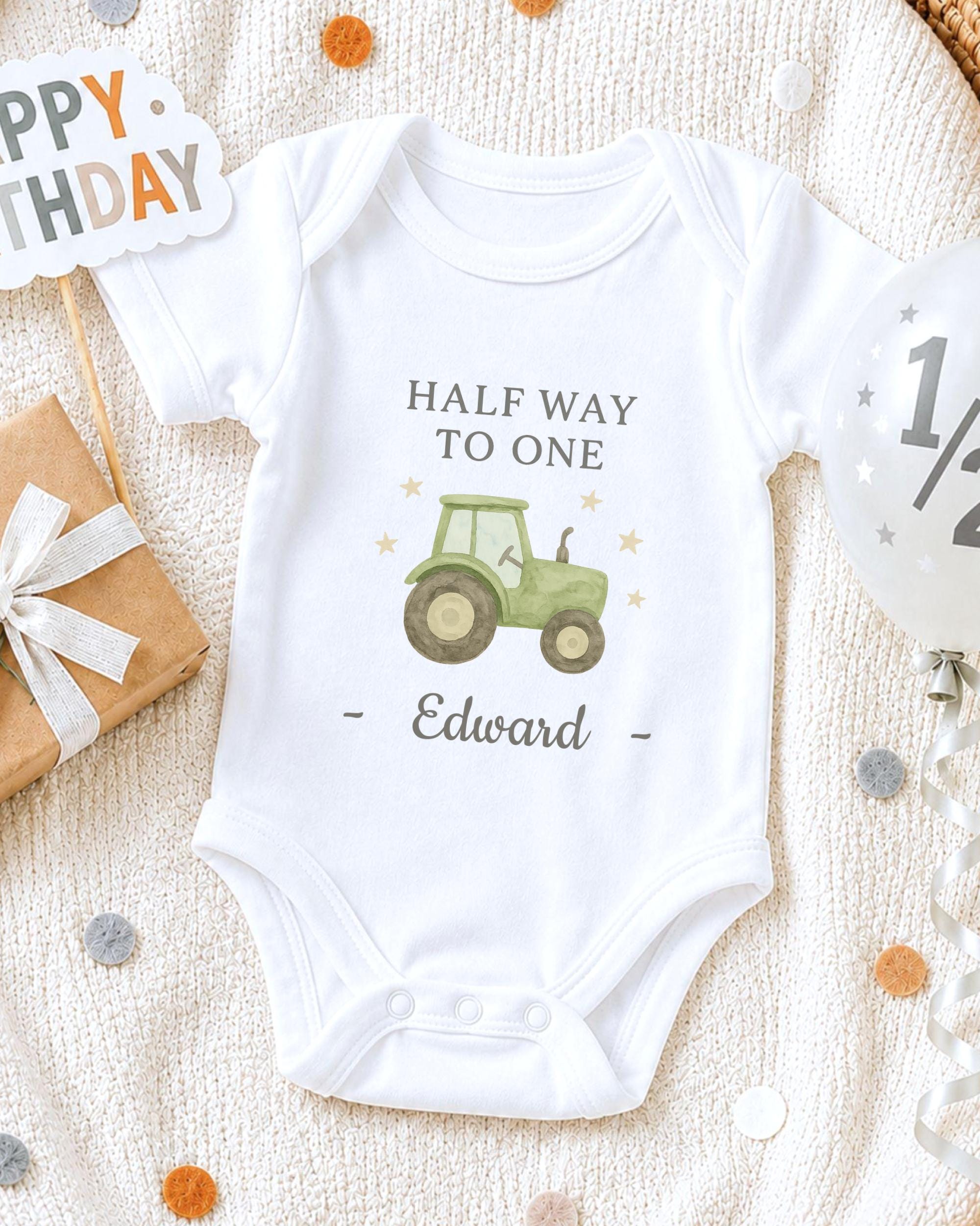 A white onesie with a tractor graphic and the text "Half Way to One Edward" printed on it, along with a birthday sign and confetti.