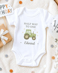 A white onesie with a tractor graphic and the text "Half Way to One Edward" printed on it, along with a birthday sign and confetti.