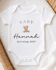 A white baby onesie with a teddy bear and the text "BABY Hannah Arriving 2027" printed on it.