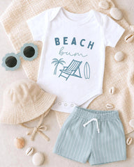 A baby's outfit consisting of a white onesie with a beach-themed design, a beige bucket hat, and striped shorts, along with a pair of sunglasses and some seashells, all laid out on a beige background.