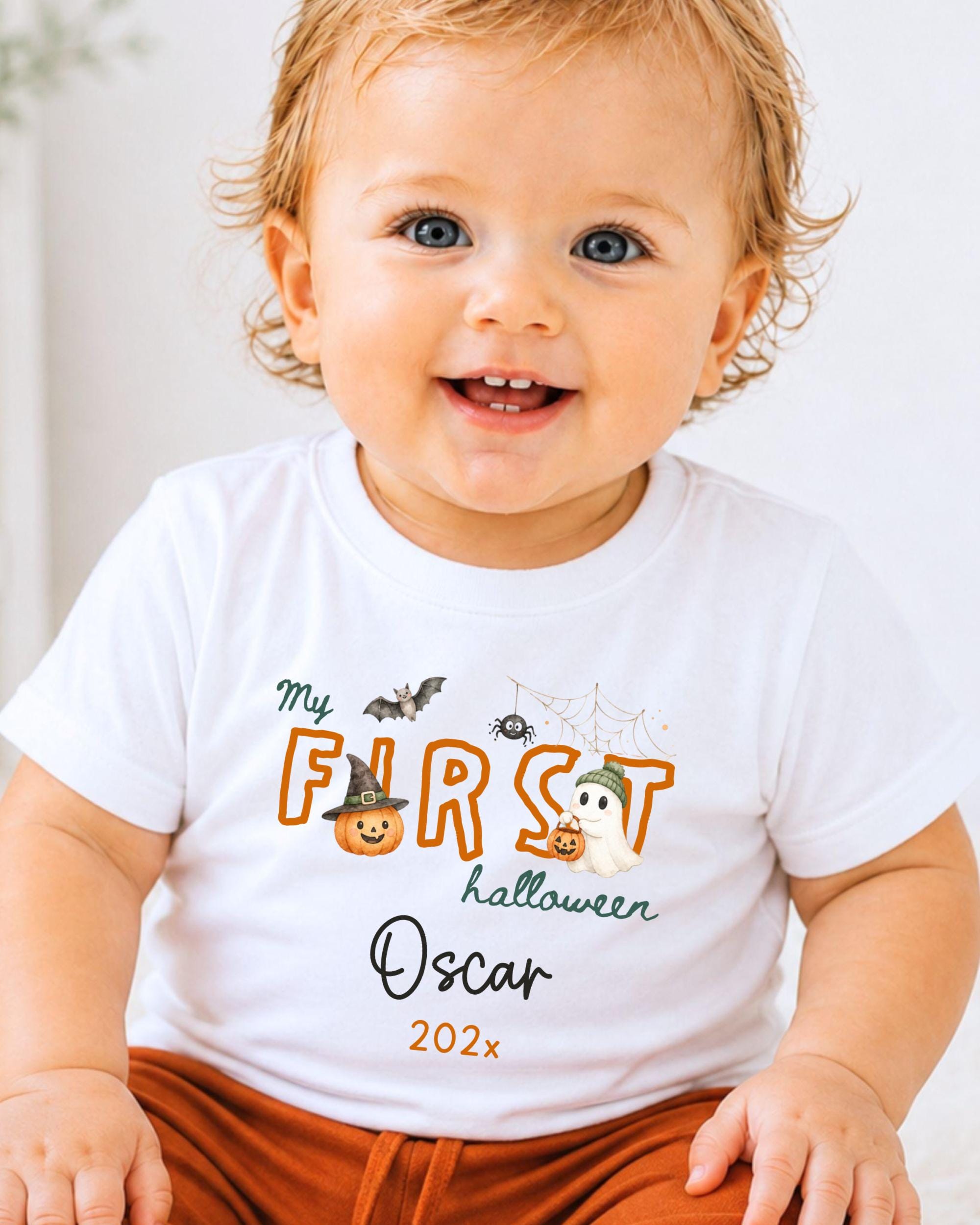 A smiling baby wearing a white t-shirt with a Halloween-themed design and text.