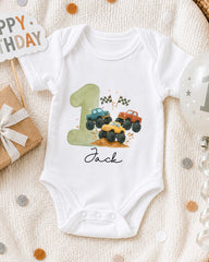 A white onesie with a graphic of a truck on it, along with a birthday sign and a balloon, all placed on a white blanket.