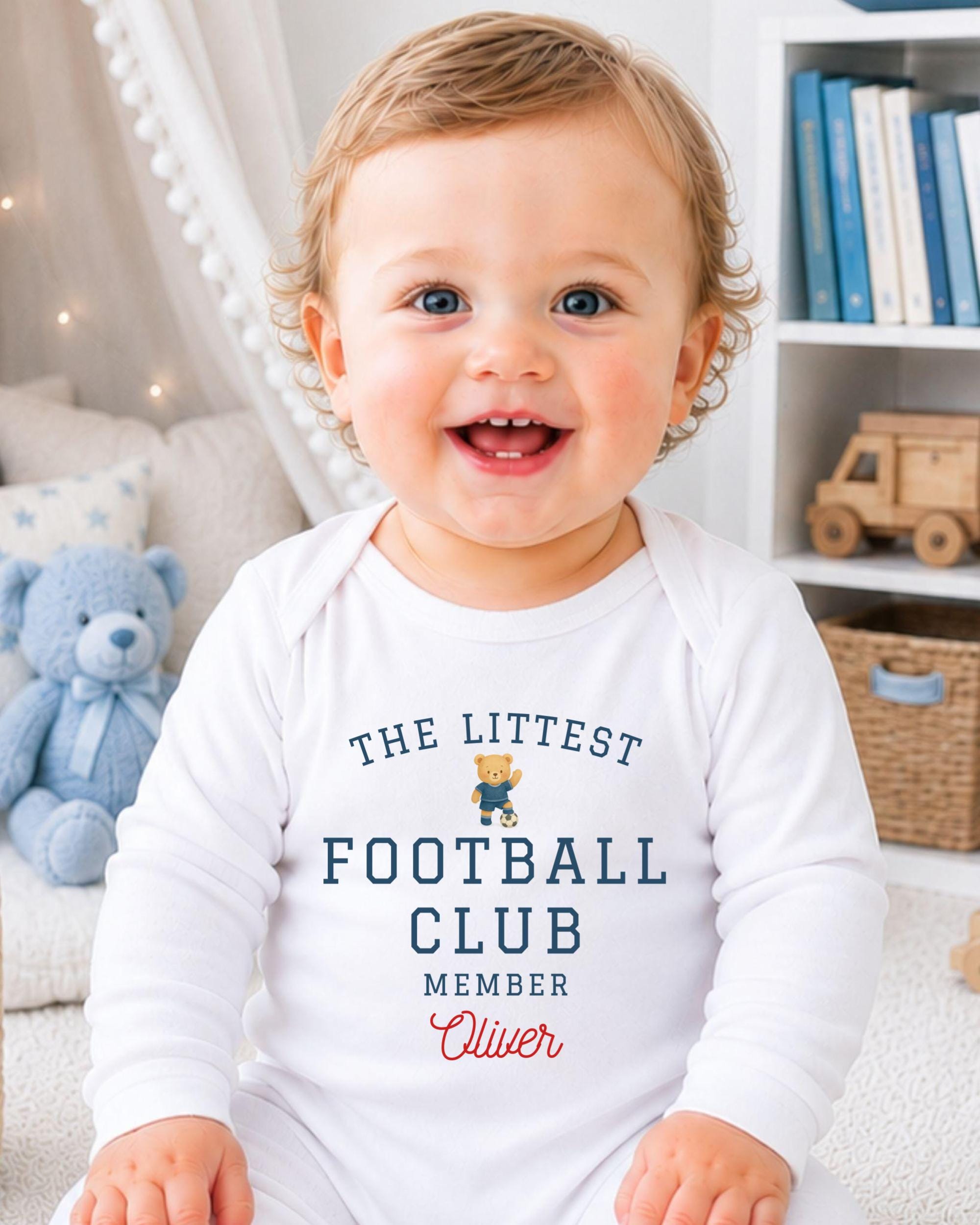 A smiling baby boy wearing a white onesie with text and a teddy bear in the background.