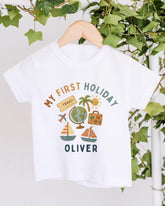 A white t-shirt with a colorful graphic design depicting various travel-related icons, including a globe, a suitcase, a palm tree, and a sailboat. The text on the t-shirt says "My first holiday Oliver".