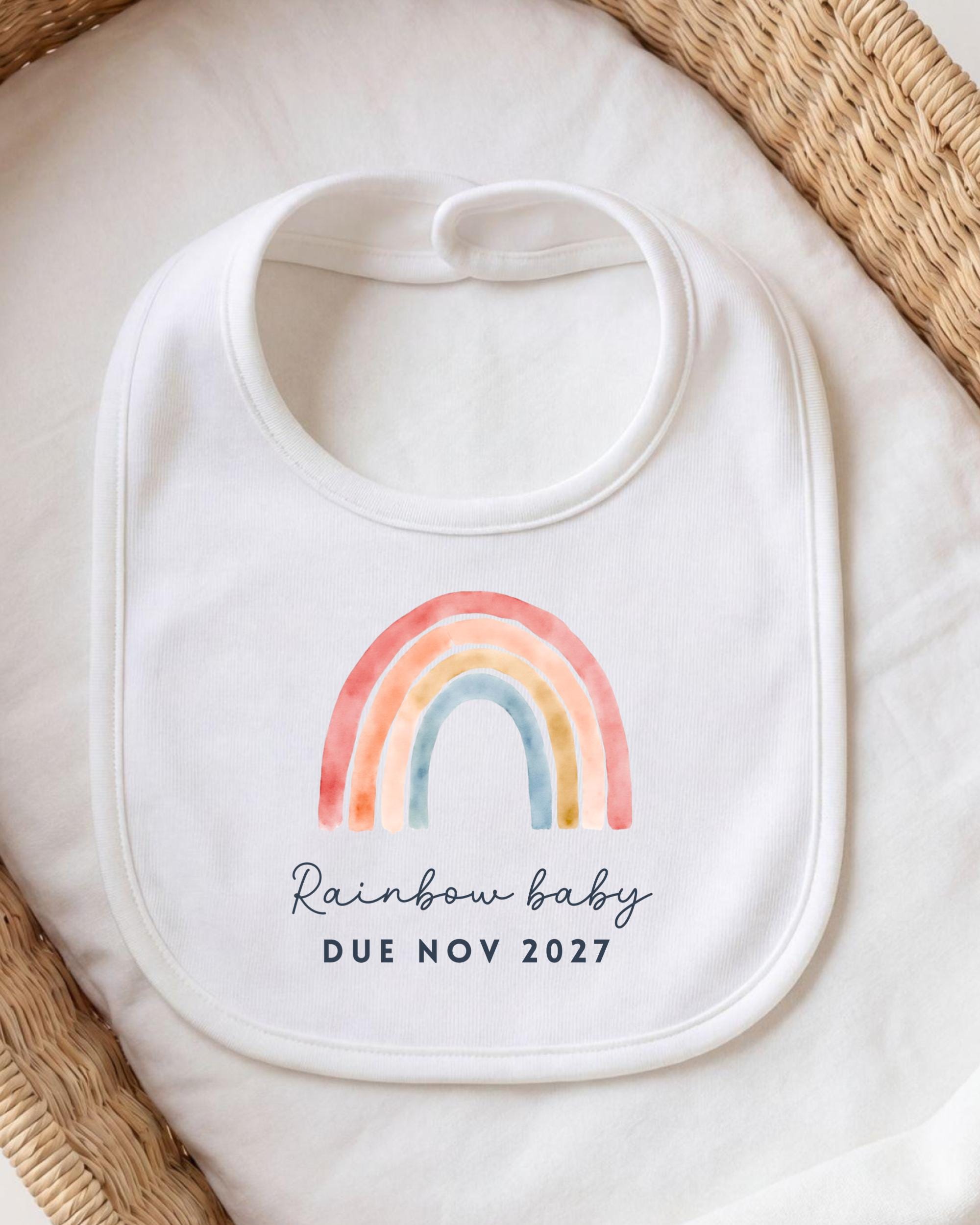 A white bib with a colorful rainbow graphic and the text "Rainbow baby due novely 2027" printed on it.