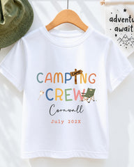 A white t-shirt with colorful text and graphics, including the words "Camping Crew" and "Cornwall" along with a camping-related image. The t-shirt is hanging on a wooden hanger, and there is a green hat and a white banner with text in the background.