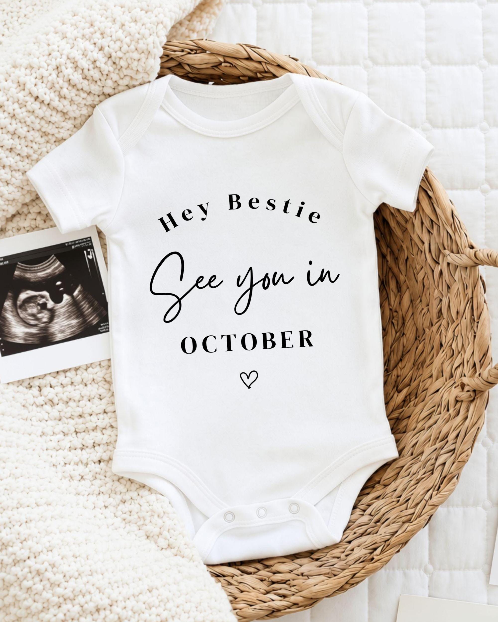A white baby onesie with the text "Hey Bestie See you in October" printed on it, placed on a woven basket.