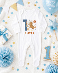 A white onesie with a dinosaur and space theme, along with a blue balloon, blue and gold confetti, and other baby-related items, all arranged on a white background.