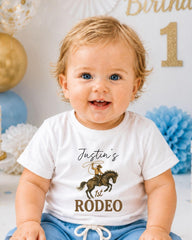 A smiling baby boy wearing a white t-shirt with a cowboy graphic and the text " Justin's 1st Rodeo" printed on it.