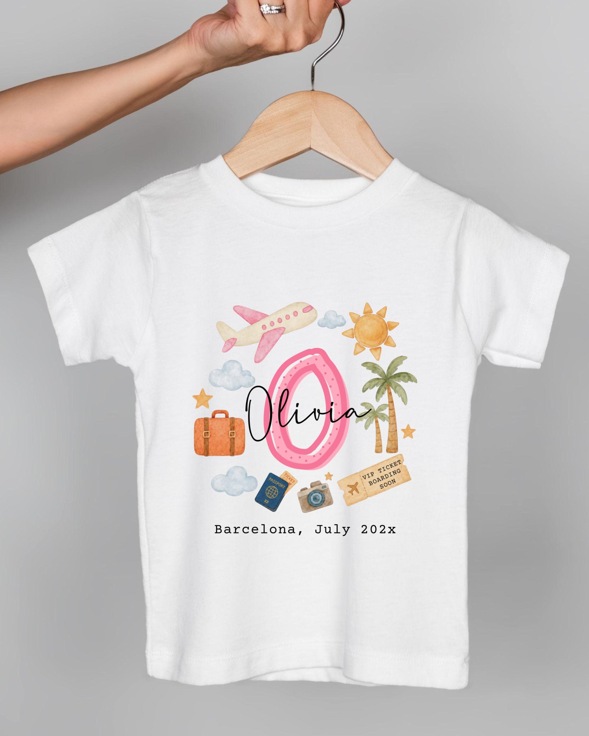 A white t-shirt with a graphic design featuring a pink number 8, a pink airplane, a suitcase, a palm tree, and various travel-related icons. The text below the design reads "Olivia Barcelona July 2023".