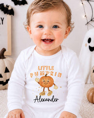 A smiling baby boy wearing a white onesie with a pumpkin character and the text "Little Pumpkin Alexander" is sitting in front of Halloween decorations.