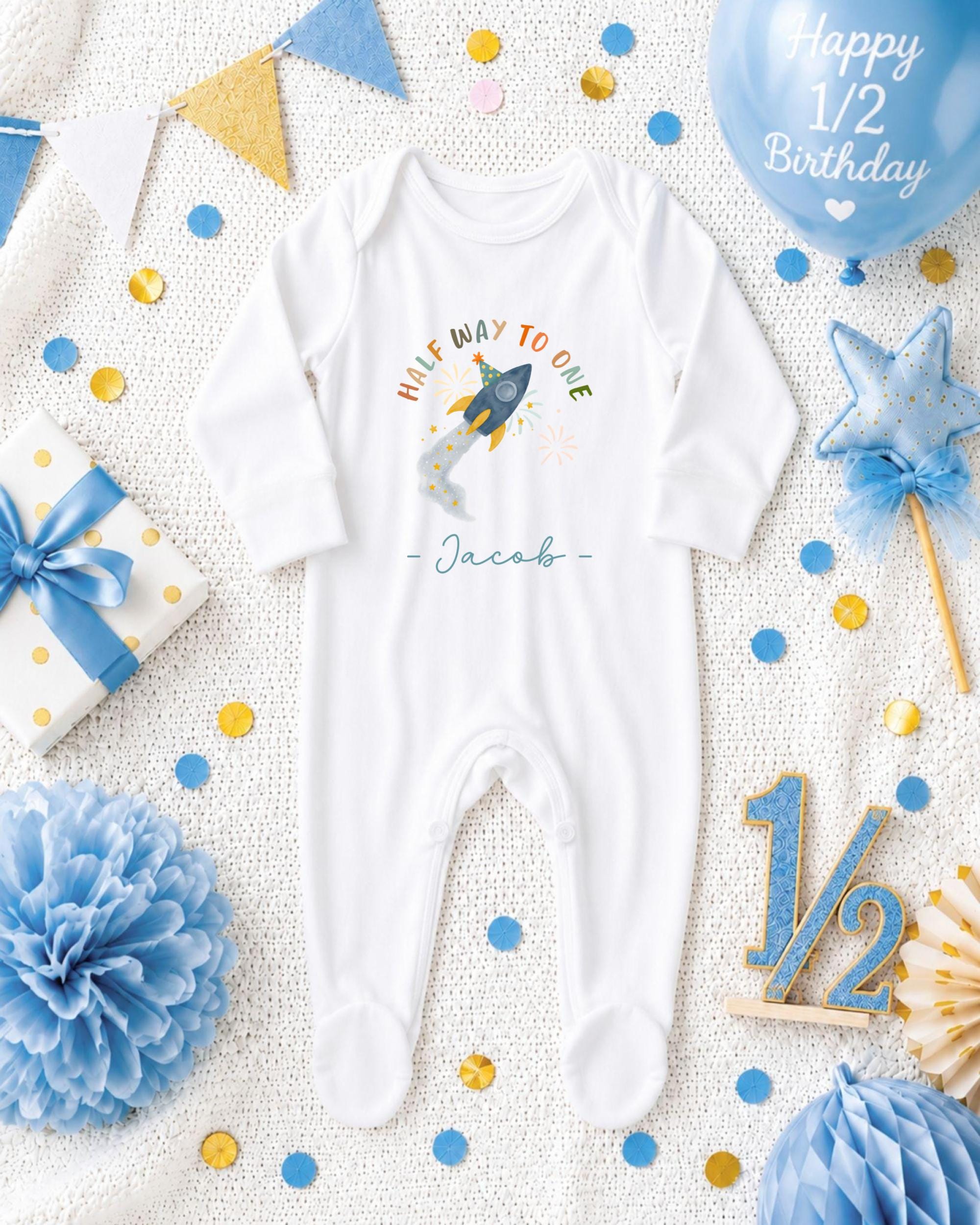 A white onesie with a rocket ship design, surrounded by blue and gold confetti, a blue balloon, and other baby-related items, all arranged on a white background.