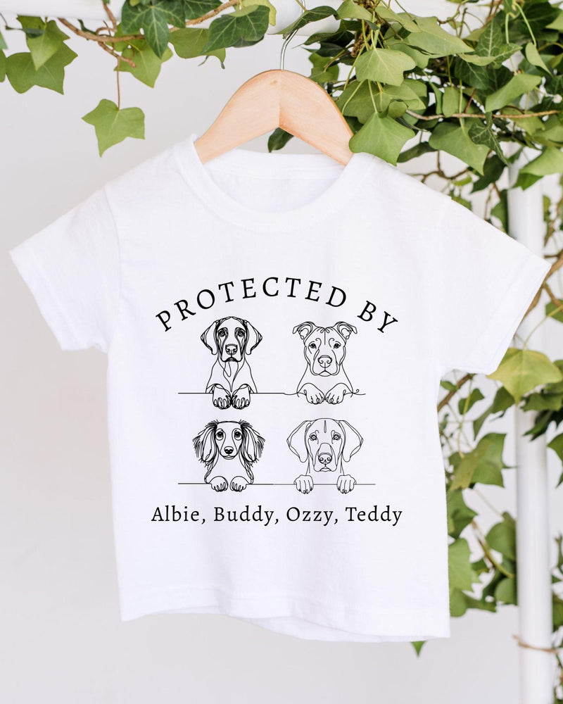A white t-shirt with a graphic design featuring four cartoon dog illustrations and the text "PROTECTED BY Albie, Buddy, Ozzy, Teddy".